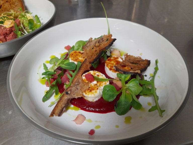 Ein kunstvoll angerichteter Teller mit Ziegenkäse, Rote-Bete-Sauce, frischem Salat und knusprigem Brot.