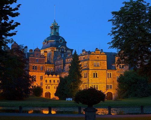 Beleuchtetes historisches Gebäude bei Dämmerung, umgeben von Bäumen und einem ruhigen Teich im Vordergrund.